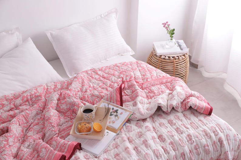 May include: A bed is made with white pillows and a pink floral patterned quilt. A wooden tray holds a cup of coffee, a pastry, and an orange slice. A small woven side table holds a vase of flowers and a book.