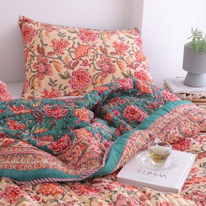 May include: A floral patterned quilt and pillow set in shades of cream, teal, and red. The quilt features a detailed floral design with a teal border. A pillow with a matching floral pattern rests on the bed. A book with the word "WHITE" is on the bed.