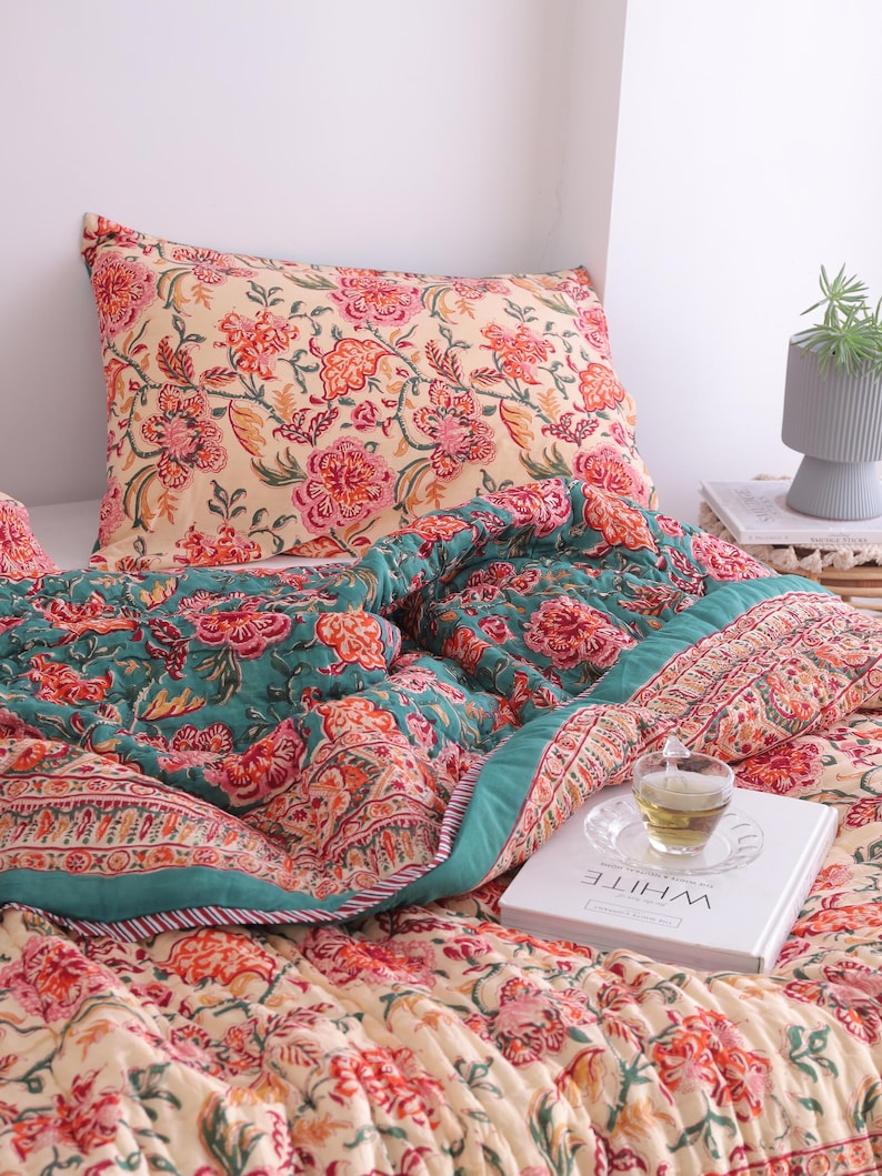 May include: A floral patterned quilt and pillow set in shades of cream, teal, and red. The quilt features a detailed floral design with a teal border. A pillow with a matching floral pattern rests on the bed. A book with the word "WHITE" is on the bed.