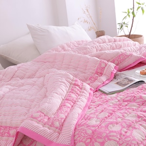 May include: A pink and white patterned quilt and matching pillows on a bed. The quilt has a floral and striped design with a solid pink border. A magazine and snacks are on the bed.