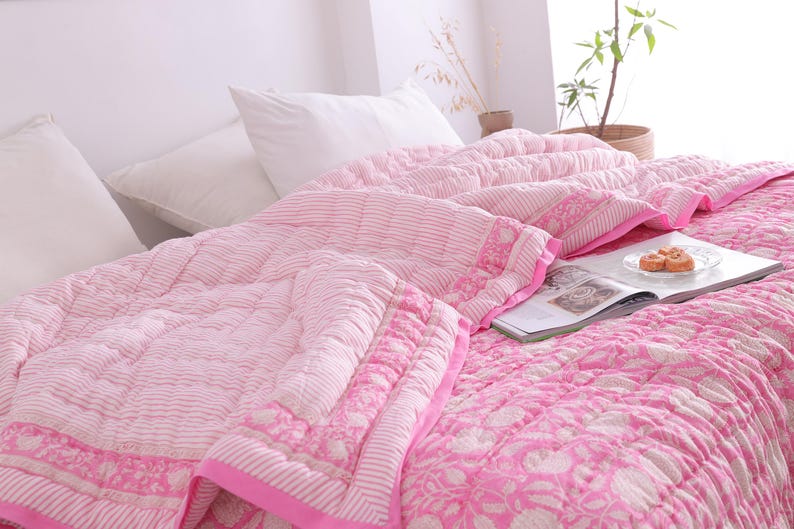 May include: A pink and white patterned quilt and matching pillows on a bed. The quilt has a floral and striped design with a solid pink border. A magazine and snacks are on the bed.