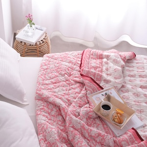 May include: A bed is covered with a pink and white patterned quilt. White pillows are on the bed. A small wicker side table holds a vase of flowers and a book. A tray with coffee and snacks sits on the quilt.