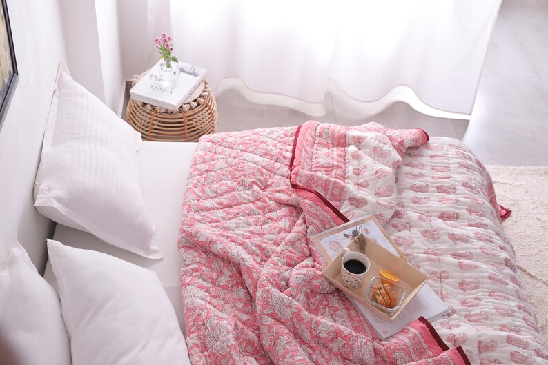 May include: A bed is covered with a pink and white patterned quilt. White pillows are on the bed. A small wicker side table holds a vase of flowers and a book. A tray with coffee and snacks sits on the quilt.