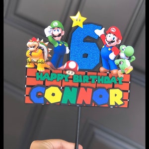 May include: A cake topper featuring Mario, Luigi, and other characters from the Super Mario Bros. video game series. The topper is decorated with a blue number six, a yellow star, and a brick wall with the words "Happy Birthday Connor".