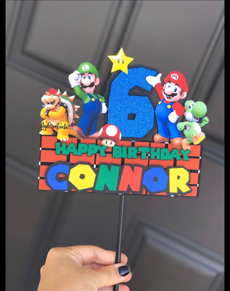 May include: A cake topper featuring Mario, Luigi, and other characters from the Super Mario Bros. video game series. The topper is decorated with a blue number six, a yellow star, and a brick wall with the words "Happy Birthday Connor".