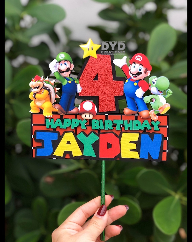 May include: A red, green, and blue cake topper with the characters Mario, Luigi, Bowser, Yoshi, and a star. The topper says "Happy Birthday Jayden" and has a red number 4 in the center.