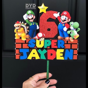 May include: A Super Mario themed cake topper with a red, green, and yellow brick background. The topper features a red number 6, the words "SUPER JAYDEN", and characters from the Super Mario video game series, including Mario, Luigi, Bowser, and Yoshi.