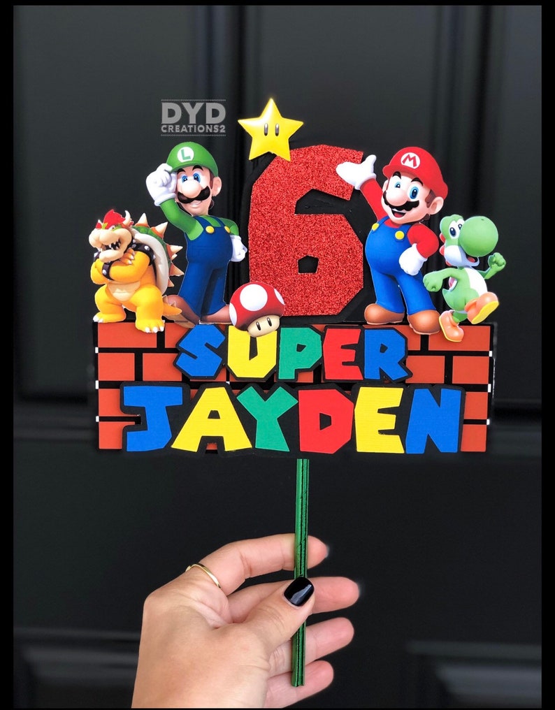 May include: A Super Mario themed cake topper with a red, green, and yellow brick background. The topper features a red number 6, the words "SUPER JAYDEN", and characters from the Super Mario video game series, including Mario, Luigi, Bowser, and Yoshi.