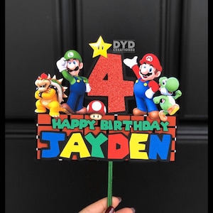 May include: A red, green, and blue cake topper with a red number 4, the words "Happy Birthday" and the name "Jayden". The topper features characters from the video game Super Mario Bros., including Mario, Luigi, Bowser, and Yoshi.