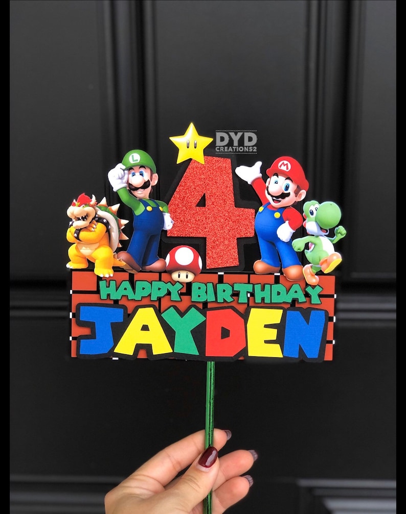 May include: A red, green, and blue cake topper with a red number 4, the words "Happy Birthday" and the name "Jayden". The topper features characters from the video game Super Mario Bros., including Mario, Luigi, Bowser, and Yoshi.