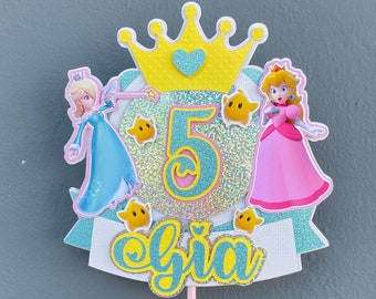 May include: A cake topper featuring Princess Peach and Rosalina from the Super Mario franchise. The topper is decorated with a yellow crown, a blue and pink glittery number 5, and the name "Gia" in yellow letters.
