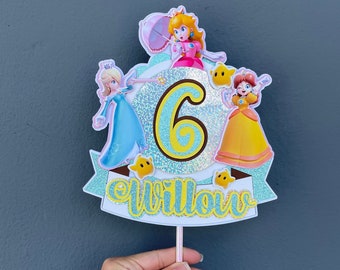 Op de afbeelding: Een taarttopper met een blauwe, gele en roze prinsesfiguur op een glinsterende achtergrond en het nummer 6. De topper bevat ook de naam "Willow" in een glinsterend lettertype.