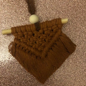 May include: A small brown macrame wall hanging with fringe, featuring a geometric pattern. It is attached to a wooden dowel with a white wooden bead at the top.