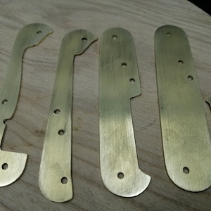 Könnte beinhalten: Fünf Messingteile für einen Messergriff, mit abgerundeten Kanten und Löchern für Nieten. Die Teile liegen auf einer Holzoberfläche.