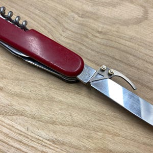 Peut inclure: Un couteau suisse rouge et argent&eacute; avec un tire-bouchon, une lame et d'autres outils. Le couteau est ouvert, montrant la lame. Le manche est rouge fonc&eacute; et les outils sont argent&eacute;s. Le couteau est sur une surface en bois.