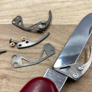 Peut inclure: Gros plan d'un couteau de poche d&eacute;mont&eacute; avec un manche rouge. La lame en acier inoxydable est partiellement ouverte, avec plusieurs petites pi&egrave;ces m&eacute;talliques, vis et &eacute;crous &eacute;parpill&eacute;s autour. Le nom de la marque du couteau est visible.