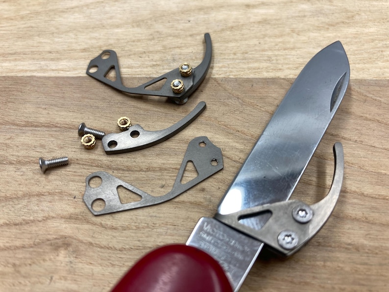 Peut inclure: Gros plan d'un couteau de poche d&eacute;mont&eacute; avec un manche rouge. La lame en acier inoxydable est partiellement ouverte, avec plusieurs petites pi&egrave;ces m&eacute;talliques, vis et &eacute;crous &eacute;parpill&eacute;s autour. Le nom de la marque du couteau est visible.