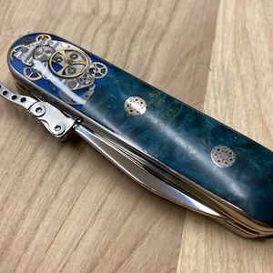 May include: A multi-tool knife with a teal and blue marbled handle. The top of the knife features a blue inlay with watch gears and gold accents. The knife has a silver metal body and a corkscrew.