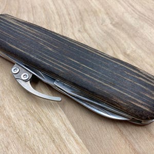 Peut inclure: Un outil multifonction avec un manche en bois fonc&eacute; et plusieurs outils en acier inoxydable. La poign&eacute;e a une forme arrondie et un clip en m&eacute;tal. L'outil est ferm&eacute; et pos&eacute; sur une surface en bois clair.
