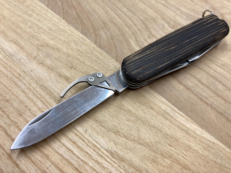 Peut inclure: Un couteau multifonction avec un manche en bois et une lame en acier inoxydable. Le couteau est &eacute;quip&eacute; d'un crochet et d'un porte-cl&eacute;s. Le manche est brun fonc&eacute; avec un grain de bois visible.