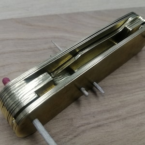 Könnte beinhalten: Ein Multifunktionswerkzeug aus Messing mit mehreren zusammen gefalteten Klingen. Das Werkzeug ist geöffnet und die Klingen sind sichtbar. Das Werkzeug befindet sich auf einer Holzoberfläche.
