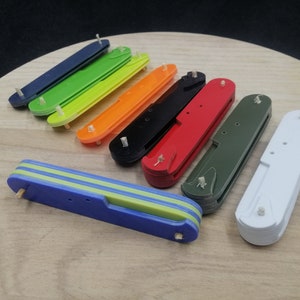 May include: A collection of eight multi-colored plastic pocket knife scales. The scales are arranged in a row on a wooden surface. The colors include blue, green, yellow, orange, black, red, green, and white.