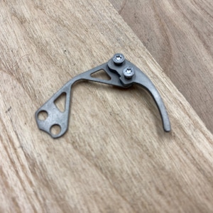 Peut inclure: Un petit composant m&eacute;tallique argent&eacute; avec un levier incurv&eacute; et deux vis. Le composant comporte une d&eacute;coupe triangulaire et deux trous &agrave; une extr&eacute;mit&eacute;. Il repose sur une surface en bois brun clair.
