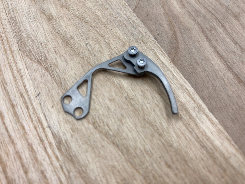 Peut inclure: Un petit composant m&eacute;tallique argent&eacute; avec un levier incurv&eacute; et deux vis. Le composant comporte une d&eacute;coupe triangulaire et deux trous &agrave; une extr&eacute;mit&eacute;. Il repose sur une surface en bois brun clair.