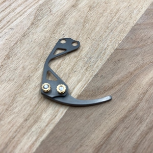 Peut inclure: Un petit composant m&eacute;tallique incurv&eacute; avec une finition gris fonc&eacute;. Il comporte deux accents dor&eacute;s et un design avec trois trous. L'objet est pos&eacute; sur une surface en bois brun clair.