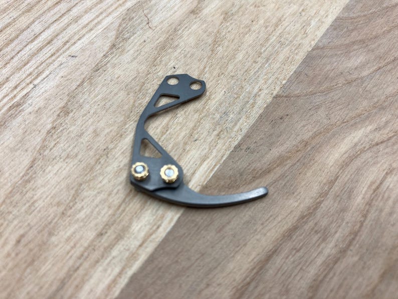 Peut inclure: Un petit composant m&eacute;tallique incurv&eacute; avec une finition gris fonc&eacute;. Il comporte deux accents dor&eacute;s et un design avec trois trous. L'objet est pos&eacute; sur une surface en bois brun clair.