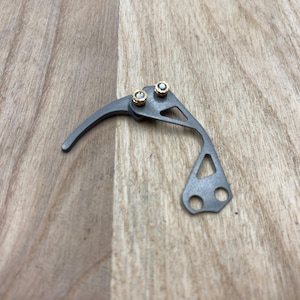 Peut inclure: Un petit composant m&eacute;canique gris m&eacute;tallique avec un bras de levier incurv&eacute; et deux rivets dor&eacute;s. Le composant pr&eacute;sente un design g&eacute;om&eacute;trique avec une ouverture triangulaire et deux trous circulaires.