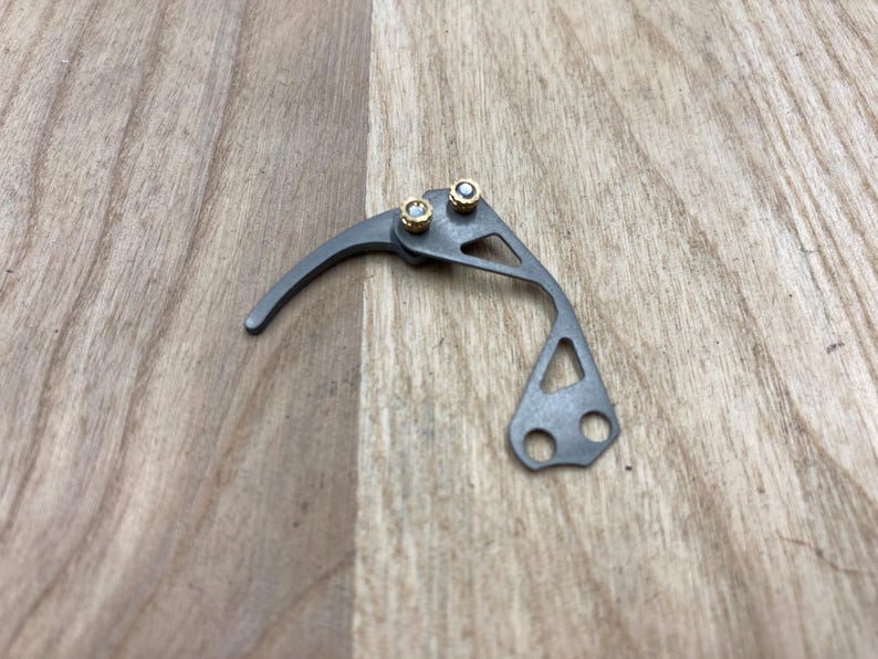 Peut inclure: Un petit composant m&eacute;canique gris m&eacute;tallique avec un bras de levier incurv&eacute; et deux rivets dor&eacute;s. Le composant pr&eacute;sente un design g&eacute;om&eacute;trique avec une ouverture triangulaire et deux trous circulaires.