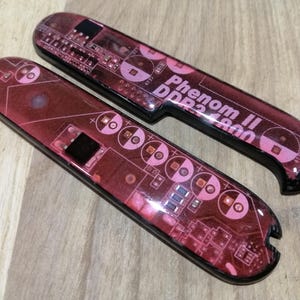 May include: Two elongated, rounded rectangular scales featuring a dark red printed circuit board pattern. The top scale displays the white text Phenom II DDR2 1800. Both pieces have a glossy finish and a small notch at one end for assembly.