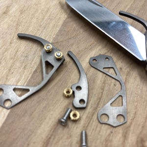 Peut inclure: Gros plan de pi&egrave;ces de couteau d&eacute;mont&eacute;es, comprenant des composants m&eacute;talliques argent&eacute;s, des vis et des &eacute;crous en laiton. La lame du couteau est partiellement visible. Les pi&egrave;ces sont dispos&eacute;es sur une surface en bois, sugg&eacute;rant une r&eacute;paration ou une personnalisation.