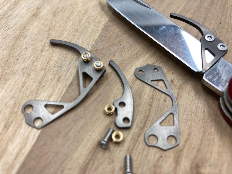 Peut inclure: Gros plan de pi&egrave;ces de couteau d&eacute;mont&eacute;es, comprenant des composants m&eacute;talliques argent&eacute;s, des vis et des &eacute;crous en laiton. La lame du couteau est partiellement visible. Les pi&egrave;ces sont dispos&eacute;es sur une surface en bois, sugg&eacute;rant une r&eacute;paration ou une personnalisation.