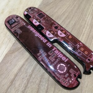 May include: Two elongated, rounded rectangular handle scales featuring a dark red and black printed circuit board pattern. The text "Designed in Taipei" is printed in white on the surface of the larger scale, which also has a notched end.