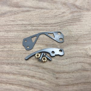 Peut inclure: Deux pi&egrave;ces m&eacute;caniques en m&eacute;tal gris avec des trous et une forme incurv&eacute;e, ainsi que de petites pi&egrave;ces en laiton et en argent. Les pi&egrave;ces sont dispos&eacute;es sur une surface en bois brun clair.