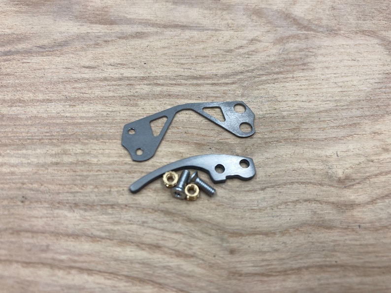 Peut inclure: Deux pi&egrave;ces m&eacute;caniques en m&eacute;tal gris avec des trous et une forme incurv&eacute;e, ainsi que de petites pi&egrave;ces en laiton et en argent. Les pi&egrave;ces sont dispos&eacute;es sur une surface en bois brun clair.