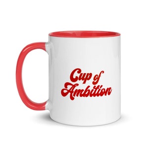 Puede incluir: Taza de cer&aacute;mica blanca con borde y asa rojos. La taza tiene el texto "Cup of Ambition" en letras rojas.