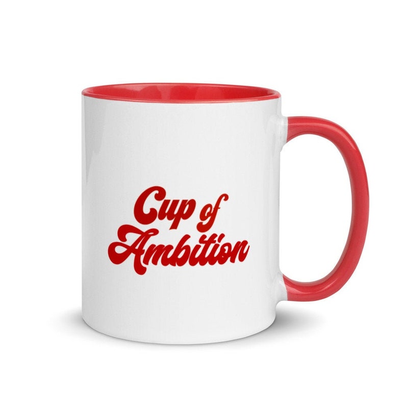 Puede incluir: Taza de cer&aacute;mica blanca con borde y asa rojos. La taza tiene el texto "Cup of Ambition" en letras rojas.