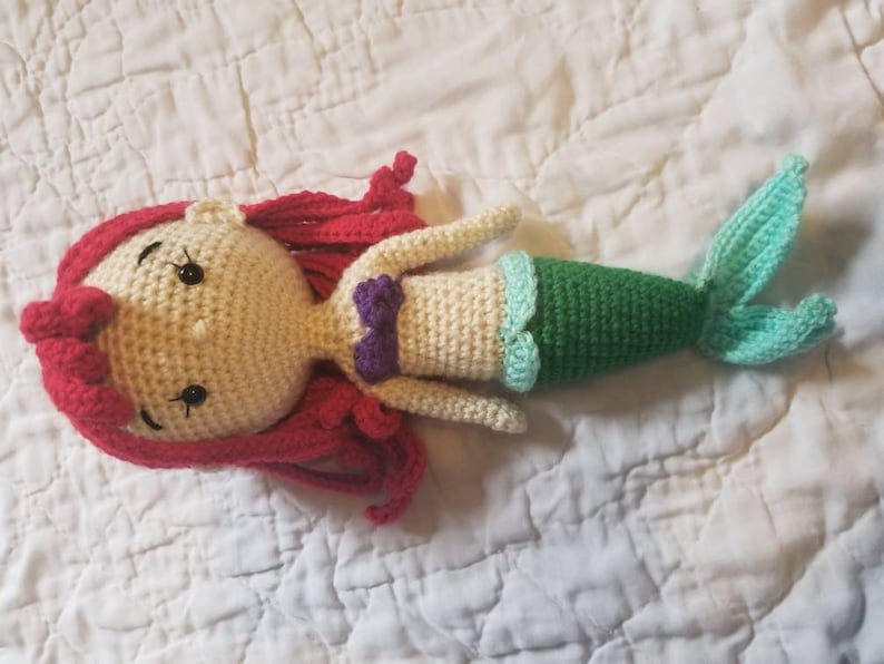 May include: A crocheted mermaid doll with red hair, a cream-colored body, a purple top, and a green and blue tail.