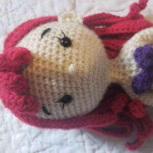May include: Crocheted doll with red hair, a purple bow, and black eyes. The doll is made of white yarn and has a crocheted dress.