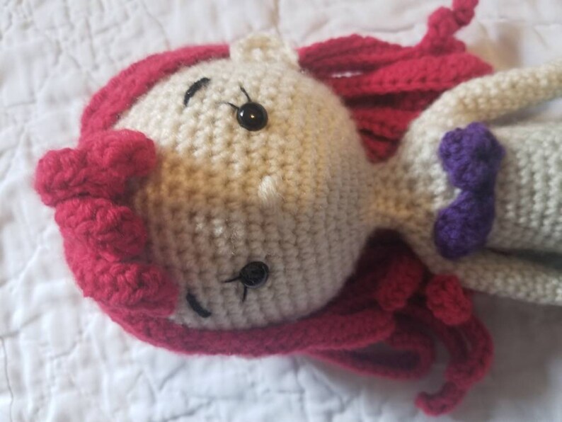 May include: Crocheted doll with red hair, a purple bow, and black eyes. The doll is made of white yarn and has a crocheted dress.