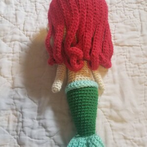 May include: A crocheted mermaid doll with red hair, a cream-colored torso, a green tail, and a light blue tail fin.