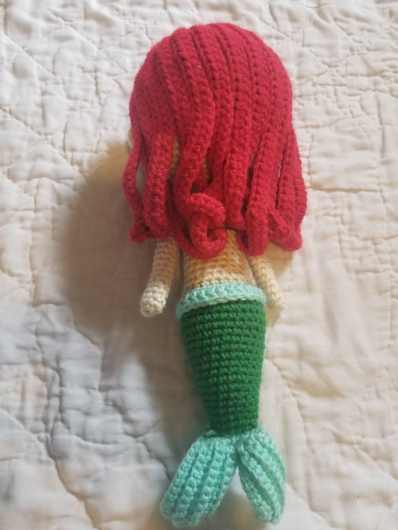May include: A crocheted mermaid doll with red hair, a cream-colored torso, a green tail, and a light blue tail fin.