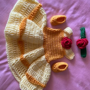 May include: A crocheted Belle costume for a baby, featuring a yellow and orange dress, a headband with a red rose, and a pair of yellow shoes.