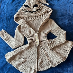 May include: A tan crochet hooded cardigan with a character face on the hood. The cardigan has long sleeves and buttons down the front. The hood features black eyes and a stitched mouth.