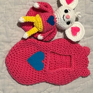 May include: A crocheted pink sleeping bag with a blue heart on the front. A white crocheted mouse with pink ears and a blue heart on its chest is lying on top of the sleeping bag.