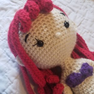 May include: A crocheted doll with red hair and a purple top. The doll has black eyes and a white face.