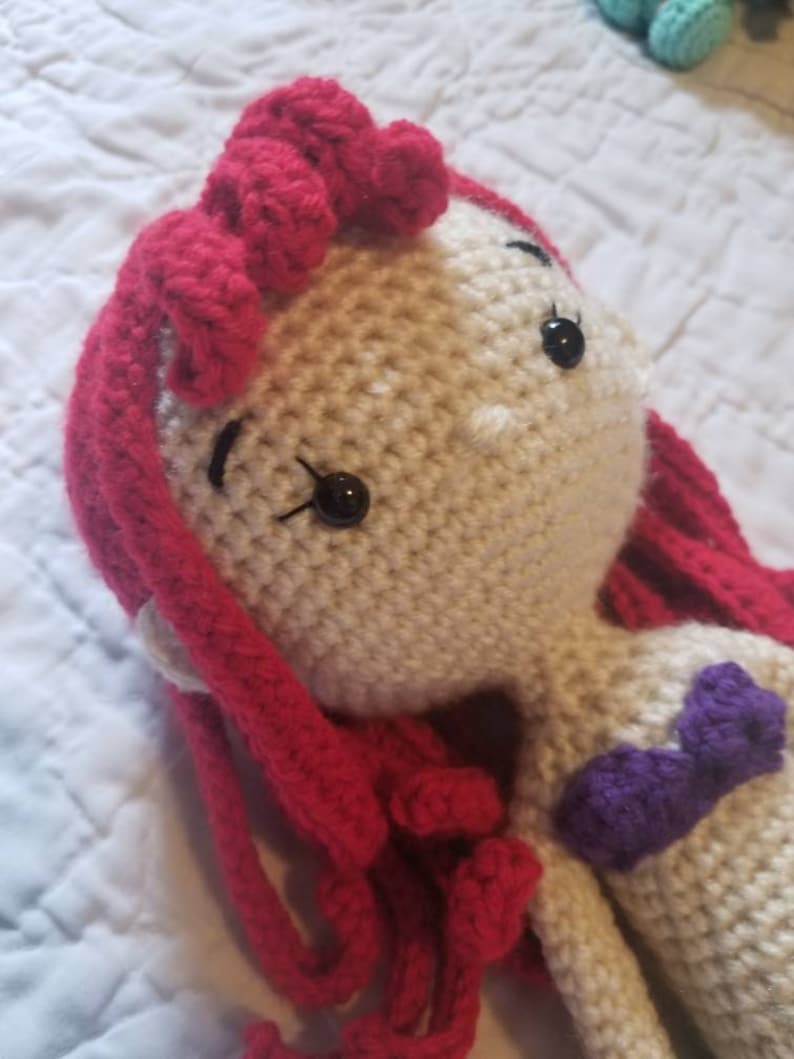 May include: A crocheted doll with red hair and a purple top. The doll has black eyes and a white face.